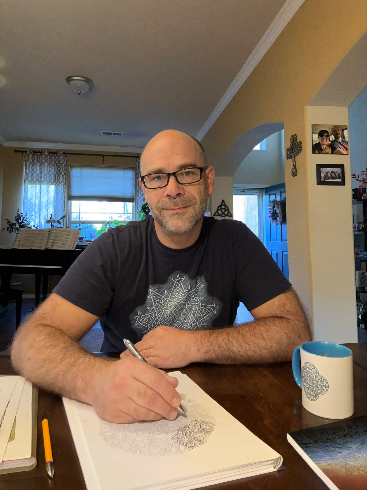 A photo of Richard Gieske, a.k.a. Xigfireon, sitting at his dining room table, working on finalizing a new Celtic knot design. Photos of his family and his piano can be seen in the background, among other things.