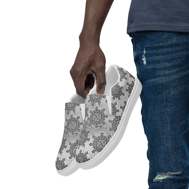 A view of a persons hand holding a pair of Xigfireon canvas slip-ons featuring the Inverted Black & White Solid Cosmos iteration of the Morning Star Fire Celtic knot pattern.