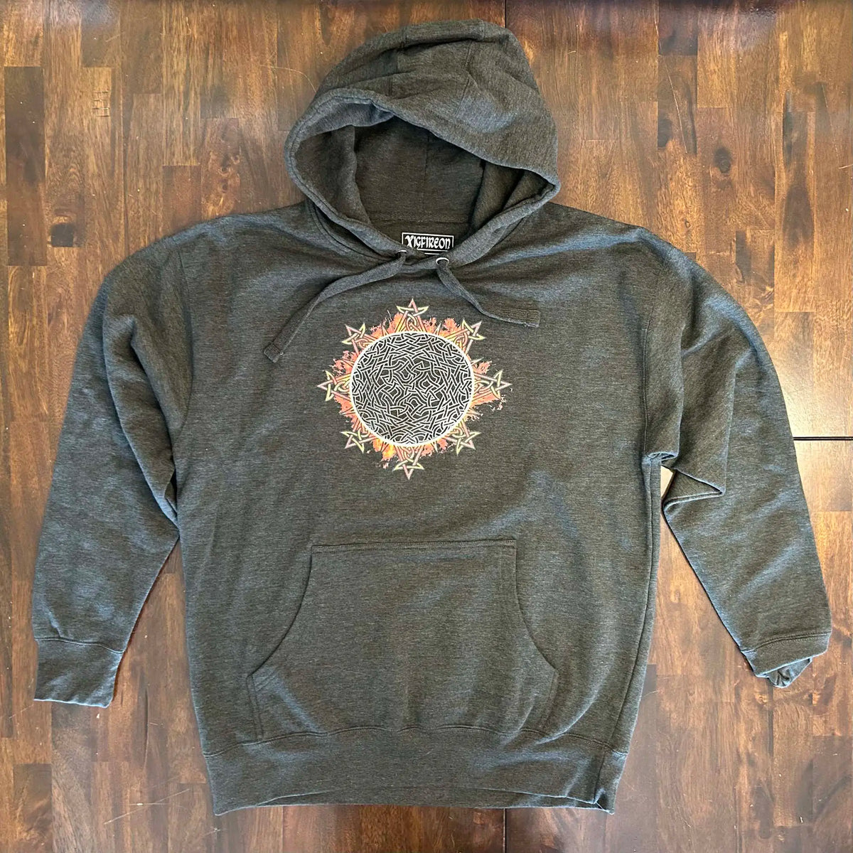 A wooden tabletop with a charcoal heather Xigfireon graphic hoodie featuring the Eclipsed iteration of the Morning Star Fire Celtic knot design, inspired by the 2023 Annular Solar Eclipse.