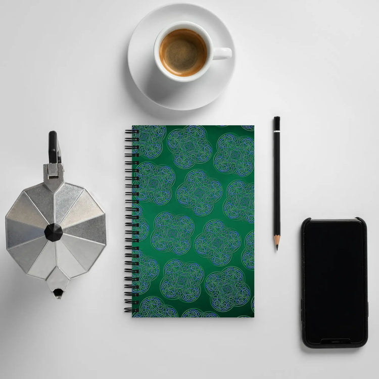A tabletop displaying a cup of espresso, creamer, a phone, a pencil, and a Xigfireon graphic spiral notebook featuring the Living Colour Forest patterned series of the Reach Of The Spirit Celtic knot design.