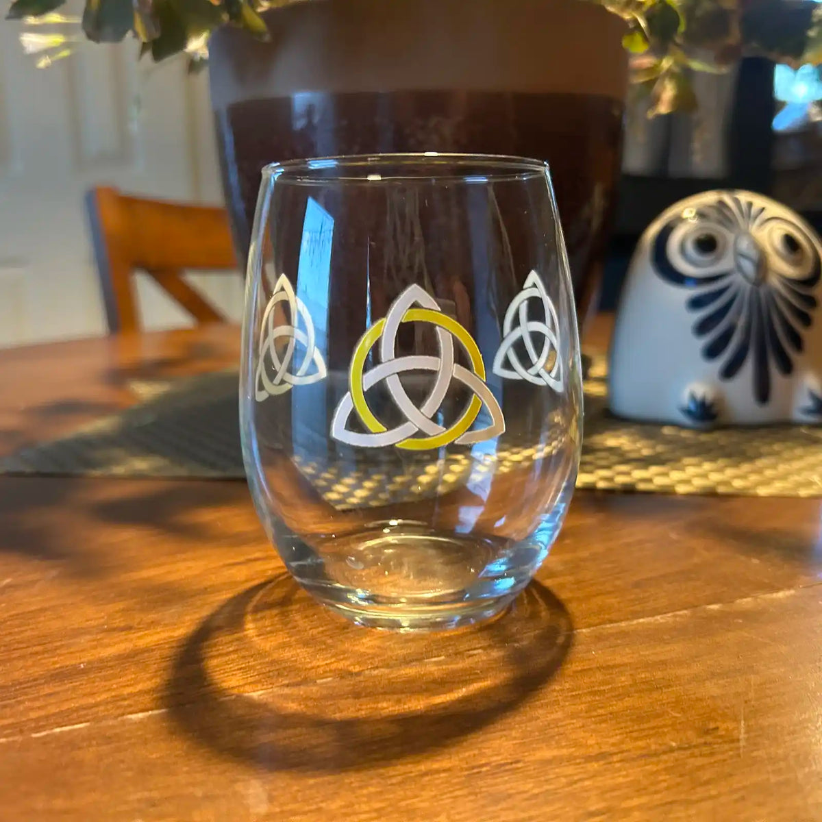 A stemless wine glass by Xigfireon with a white and yellow Trinity Knot (triquetra) design sitting on a wooden table, with a plant and decorative owl in the background.