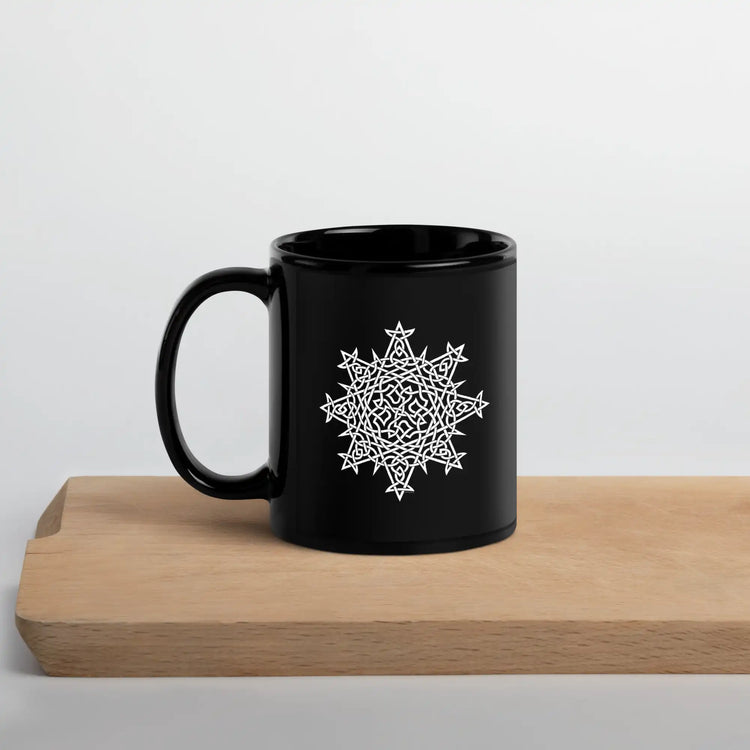 A Xigfireon black glossy 11oz mug featuring the Black & White Solid iteration of the `Morning Star Fire` Celtic knot design sitting on a cutting board. This Celtic knot symbolizes Father Sun.