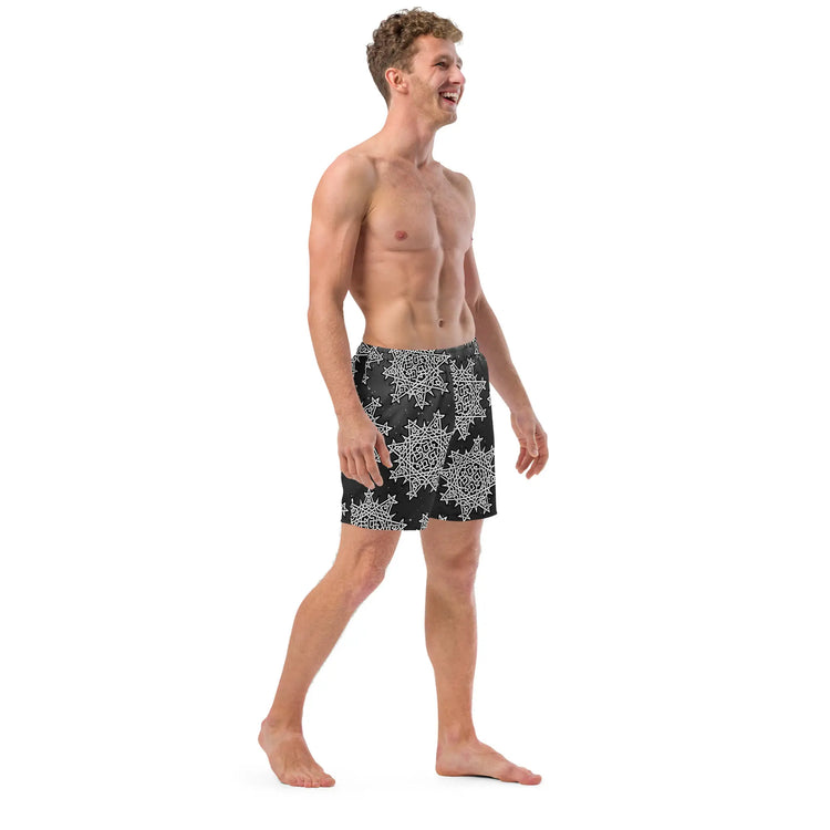 A young man wearing a pair of Xigfireon graphic swim trunks featuring the Black & White Solid Cosmos patterned series of the `Morning Star Fire` Celtic knot design.