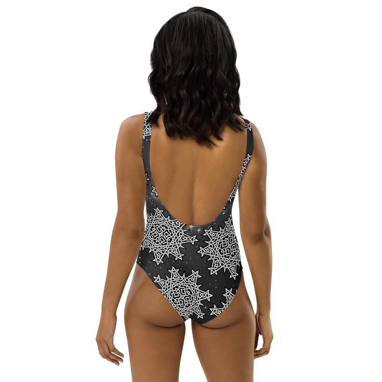 A woman, viewed from behind, wearing a Xigfireon graphic swimsuit featuring the Black & White Solid Cosmos patterned series of the `Morning Star Fire` Celtic knot design.