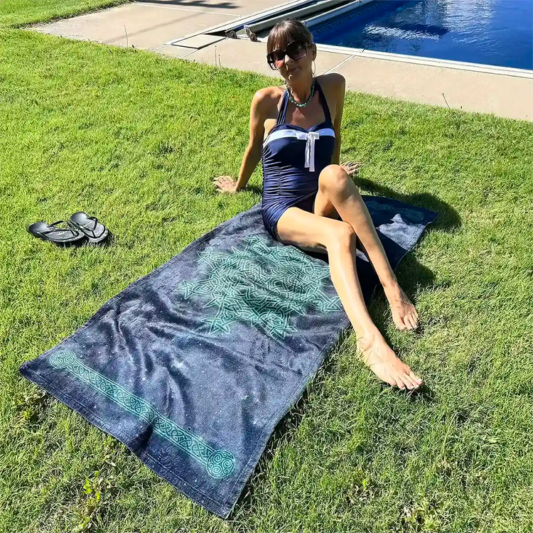 A young lady shown sunbathing near a pool on a Xigfireon beach towel featuring the Celestial iteration of the `Morning Star Fire` Celtic knot.