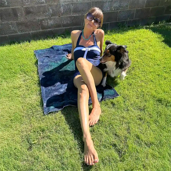 A young lady with her dog shown sunbathing on a Xigfireon beach towel featuring the Celestial iteration of the `Morning Star Fire` Celtic knot.