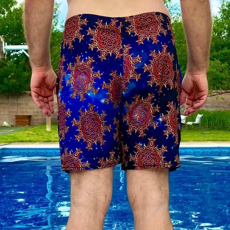 A man standing near a pool, wearing a pair of Xigfireon graphic swim trunks featuring the Fire Colour Cosmos (small) patterned series of the `Morning Star Fire` Celtic knot design.
