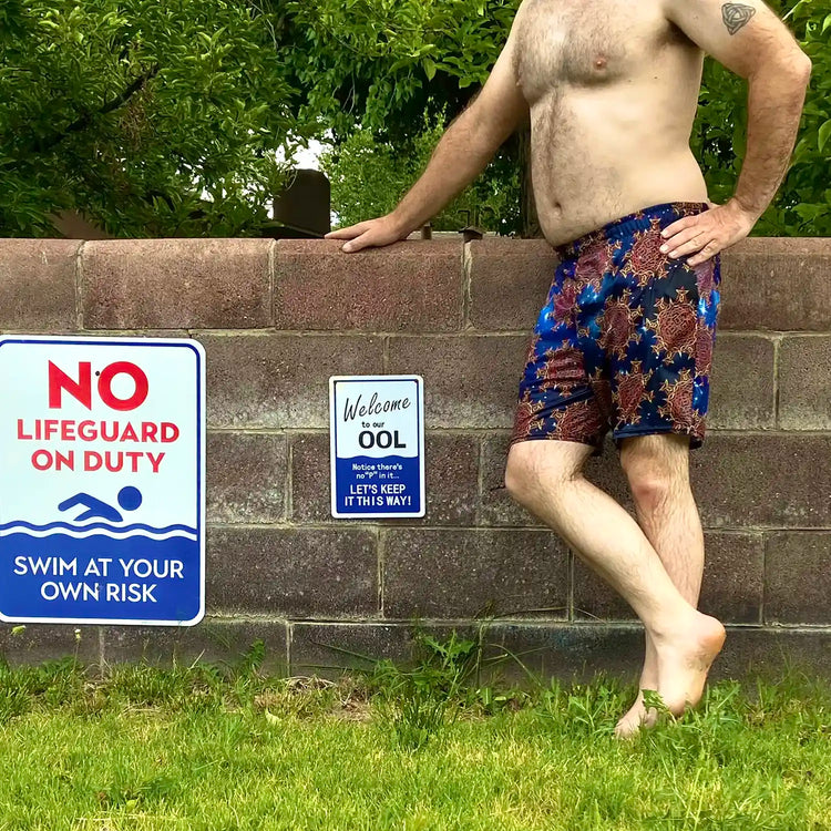 A man standing next to a wall, wearing a pair of Xigfireon graphic swim trunks featuring the Fire Colour Cosmos (small) patterned series of the `Morning Star Fire` Celtic knot design.