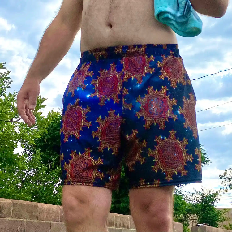 A man, viewed from the front, wearing a pair of Xigfireon graphic swim trunks featuring the Fire Colour Cosmos (small) patterned series of the `Morning Star Fire` Celtic knot design.