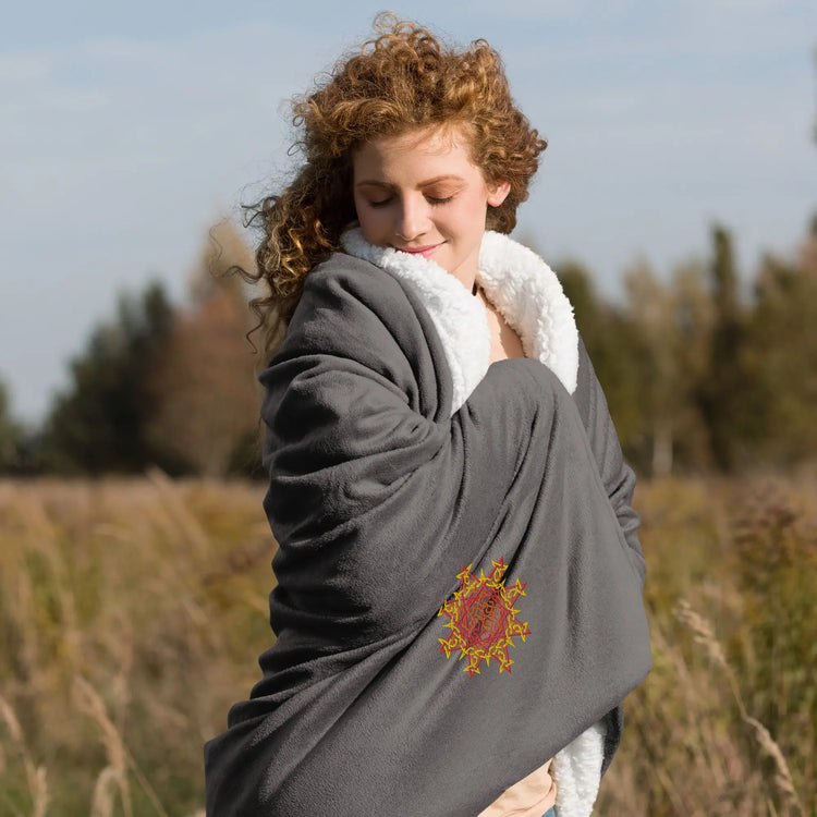 A woman in a field wrapping in a heather grey Xigfireon sherpa blanket embroidered with the Fire Colour iteration of the `Morning Star Fire` Celtic knot design.
