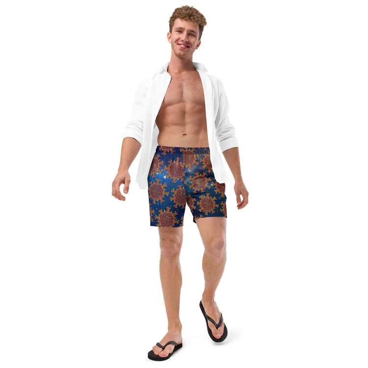 A young man wearing a pair of Xigfireon graphic swim trunks featuring the Fire Colour Cosmos (small) patterned series of the `Morning Star Fire` Celtic knot design.