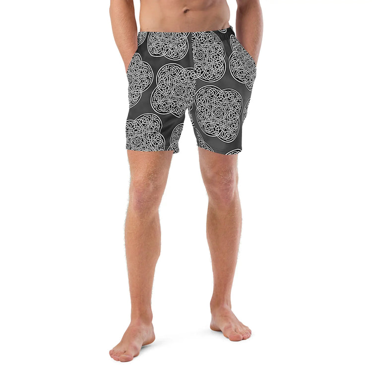 A young man wearing a pair of Xigfireon graphic swim trunks featuring the Black & White Solid Forest patterned series of the `Reach Of The Spirit` Celtic knot design.