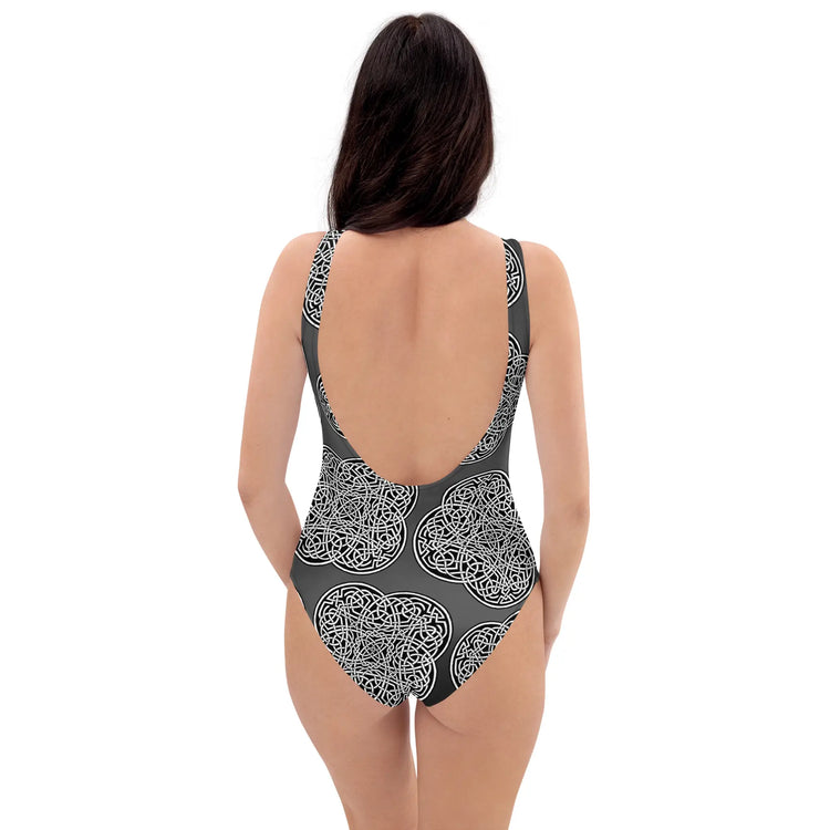A woman, viewed from behind, wearing a Xigfireon graphic swimsuit featuring the Black & White Solid Forest patterned series of the `Reach Of The Spirit` Celtic knot design.