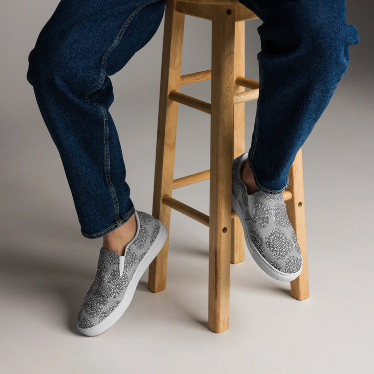 A view of a persons feet wearing a pair of Xigfireon canvas slip-ons featuring the Inverted Black & White Solid iteration of the `Reach of The Spirit` Celtic knot pattern.