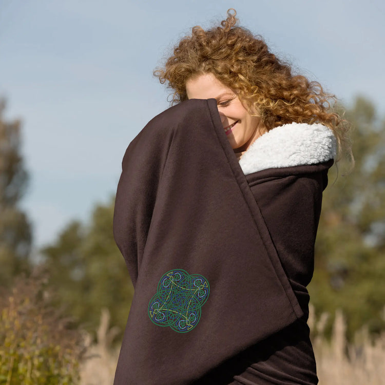 A woman in a field wrapped in a fireside brown Xigfireon sherpa blanket embroidered with the Living Colour iteration of the `Reach Of The Spirit` Celtic knot design.