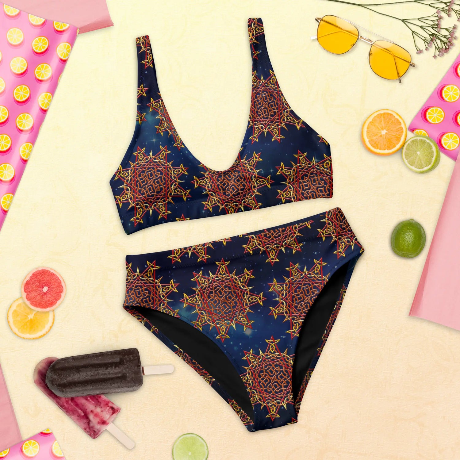 A pair of sunglasses, a couple popsicles, and some orange slices displayed with a Xigfireon graphic bikini featuring the Fire Colour Cosmos patterned series of the Morning Star Fire Celtic knot design.