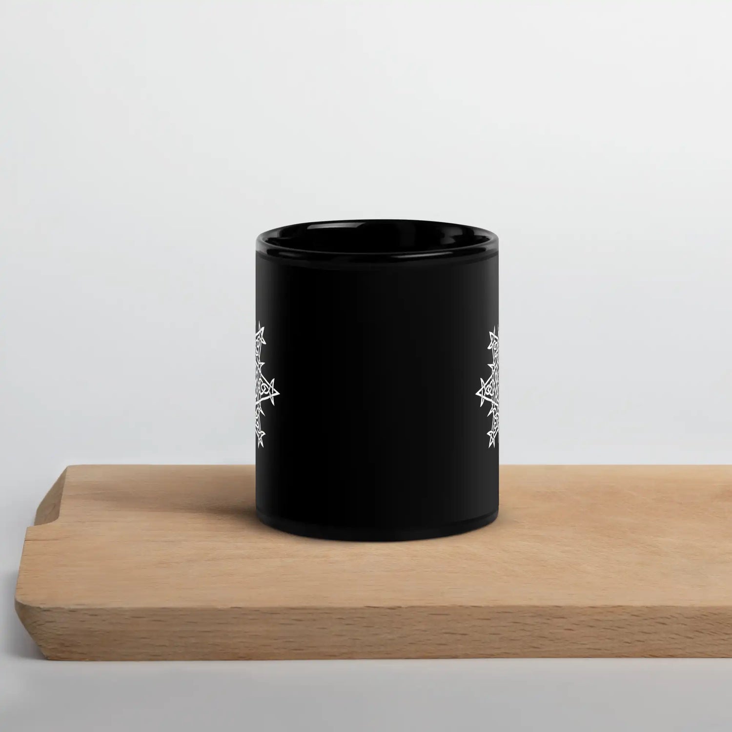 A Xigfireon black glossy 11oz mug, featuring the Black & White Solid iteration of the Morning Star Fire Celtic knot design, sitting on a cutting board. This Celtic knot symbolizes Father Sun.
