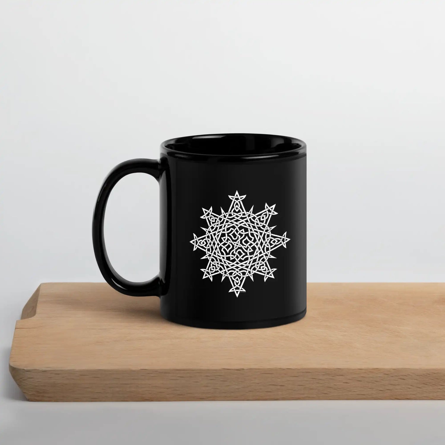 A Xigfireon black glossy 11oz mug featuring the Black & White Solid iteration of the Morning Star Fire Celtic knot design sitting on a cutting board. This Celtic knot symbolizes Father Sun.
