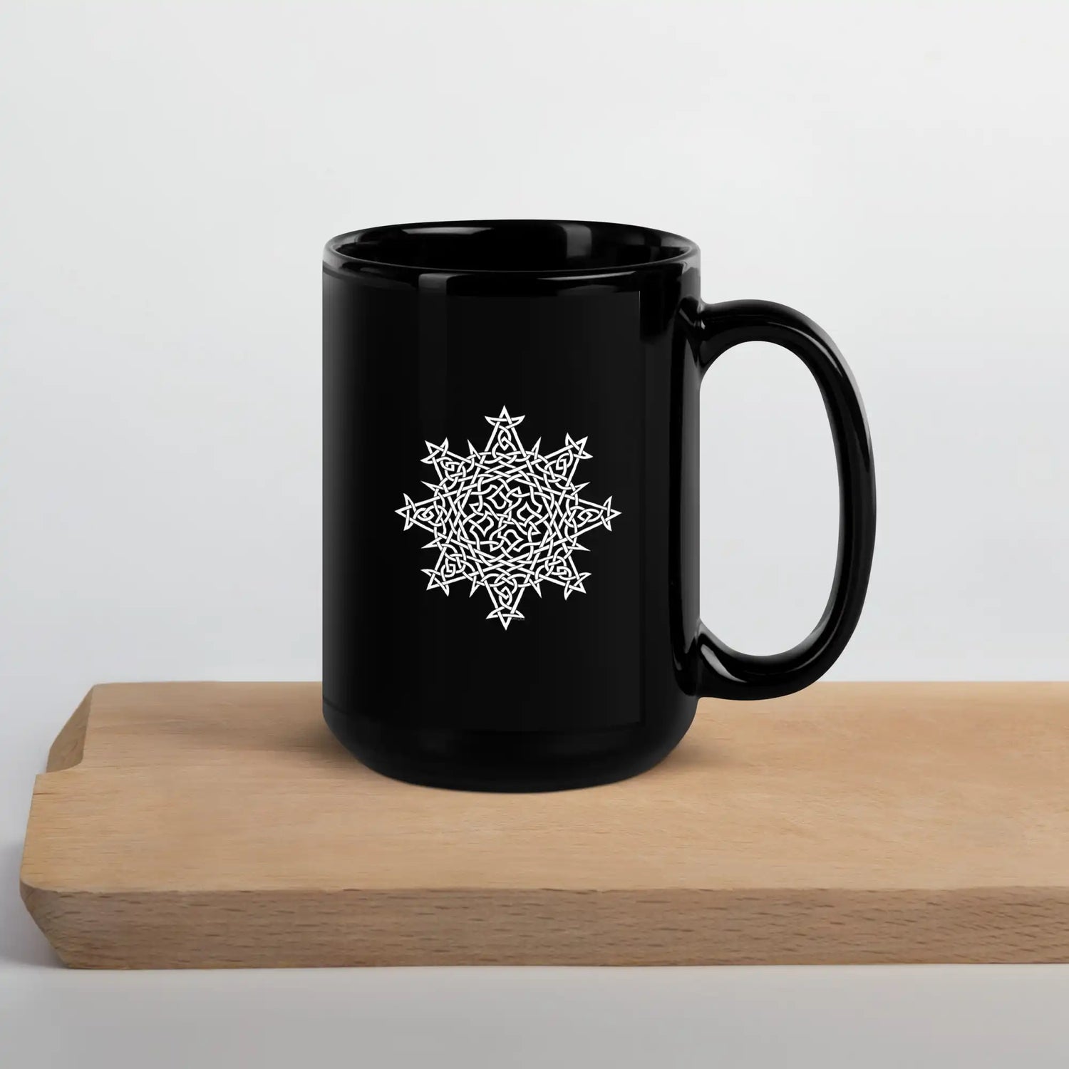 A cutting board beneath a Xigfireon black glossy 15oz mug featuring the Black & White Solid iteration of the Morning Star Fire Celtic knot design. This Celtic knot symbolizes Father Sun.