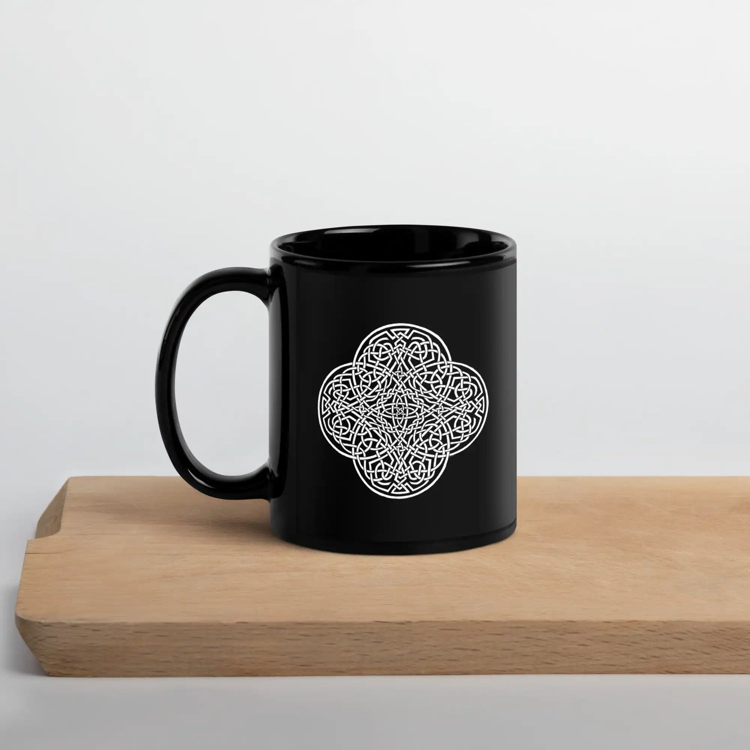 A cutting board beneath a Xigfireon black glossy 11oz mug featuring the Black & White Solid iteration of the Reach Of The Spirit Celtic knot design. This Celtic knot symbolizes Mother Earth.