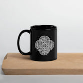 A cutting board beneath a Xigfireon black glossy 11oz mug featuring the Black & White Solid iteration of the Reach Of The Spirit Celtic knot design. This Celtic knot symbolizes Mother Earth.