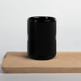 A cutting board beneath a Xigfireon black glossy 15oz mug featuring the Black & White Solid iteration of the Reach Of The Spirit Celtic knot design. This Celtic knot symbolizes Mother Earth.