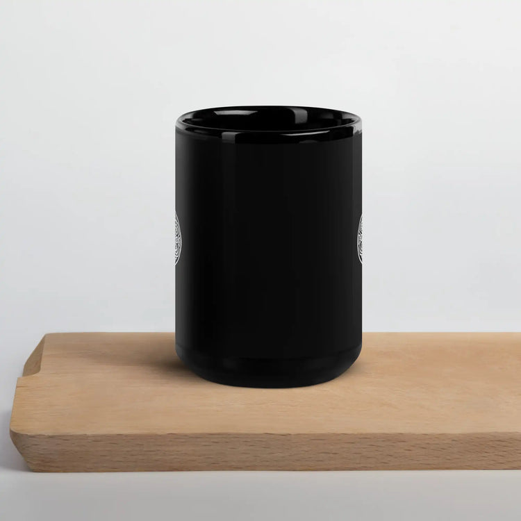 A cutting board beneath a Xigfireon black glossy 15oz mug featuring the Black & White Solid iteration of the Reach Of The Spirit Celtic knot design. This Celtic knot symbolizes Mother Earth.