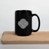 A cutting board beneath a Xigfireon black glossy 15oz mug featuring the Black & White Solid iteration of the Reach Of The Spirit Celtic knot design. This Celtic knot symbolizes Mother Earth.