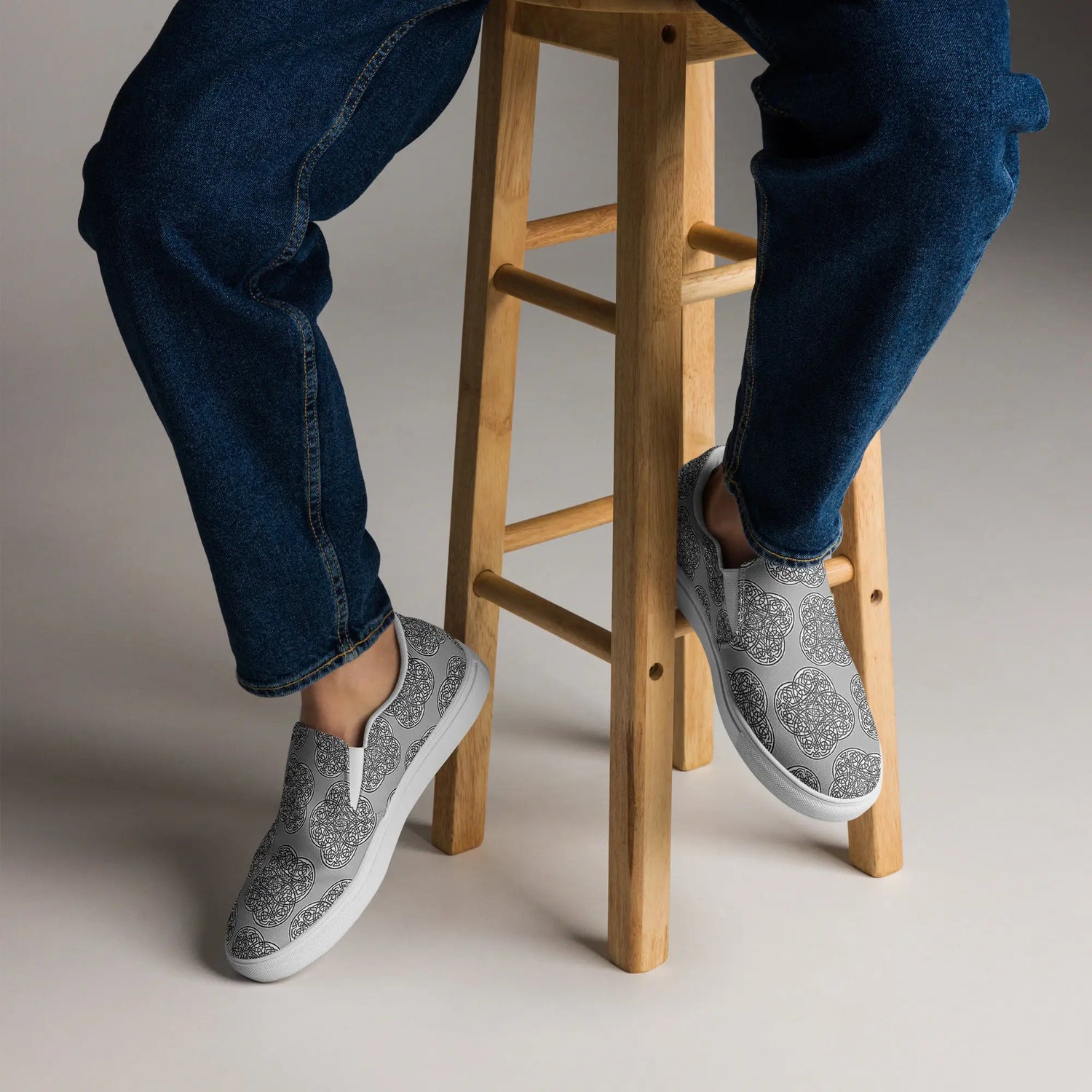 A view of a persons feet wearing a pair of Xigfireon canvas slip-ons featuring the Inverted Black & White Solid iteration of the Reach of The Spirit Celtic knot pattern.