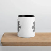 A cutting board displaying a Xigfireon white ceramic 11oz mug featuring the Black & White Solid iteration of the Morning Star Fire Celtic knot design, with black handle and inside. This Celtic knot symbolizes Father Sun.