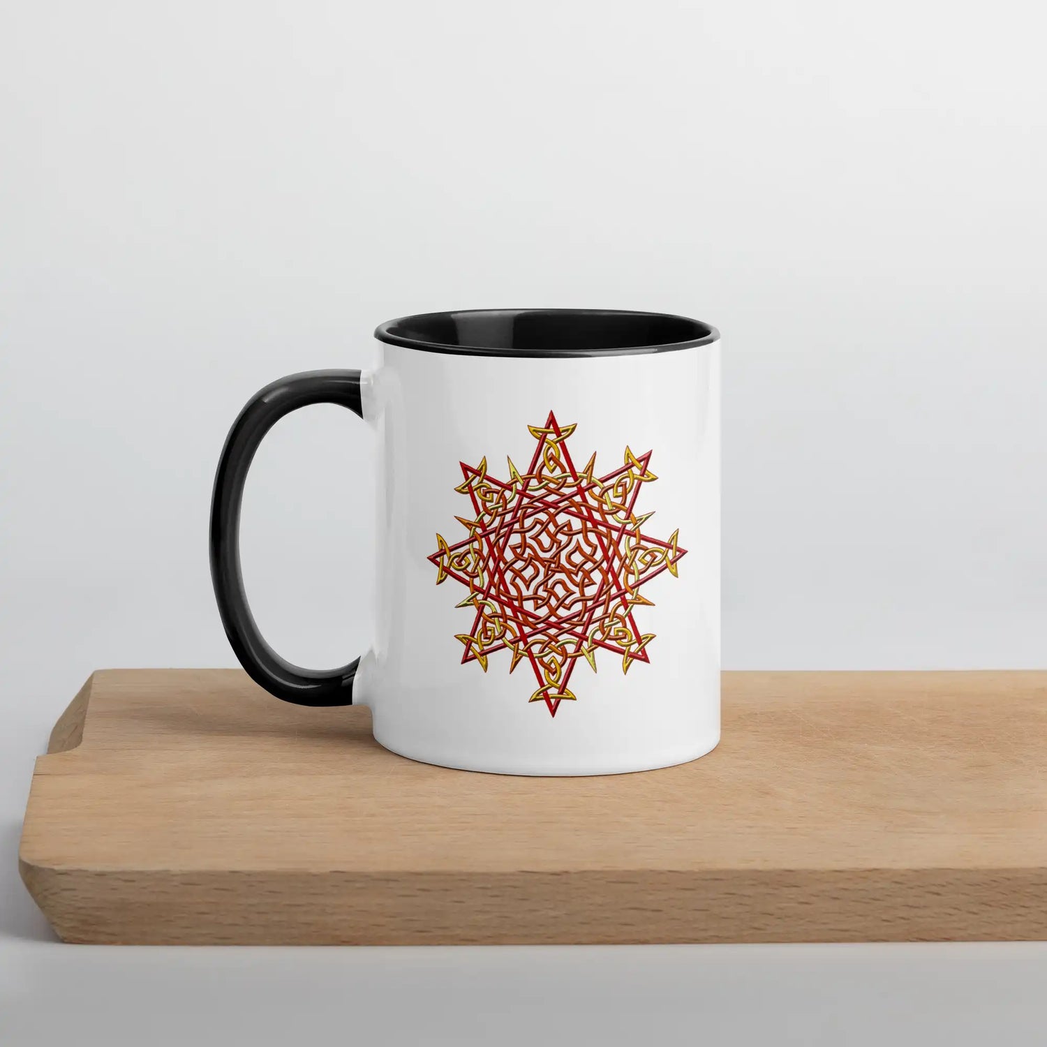A cutting board displaying a Xigfireon white ceramic 11oz mug featuring the Fire Colour iteration of the Morning Star Fire Celtic knot design, with black handle and inside. This Celtic knot symbolizes Father Sun.