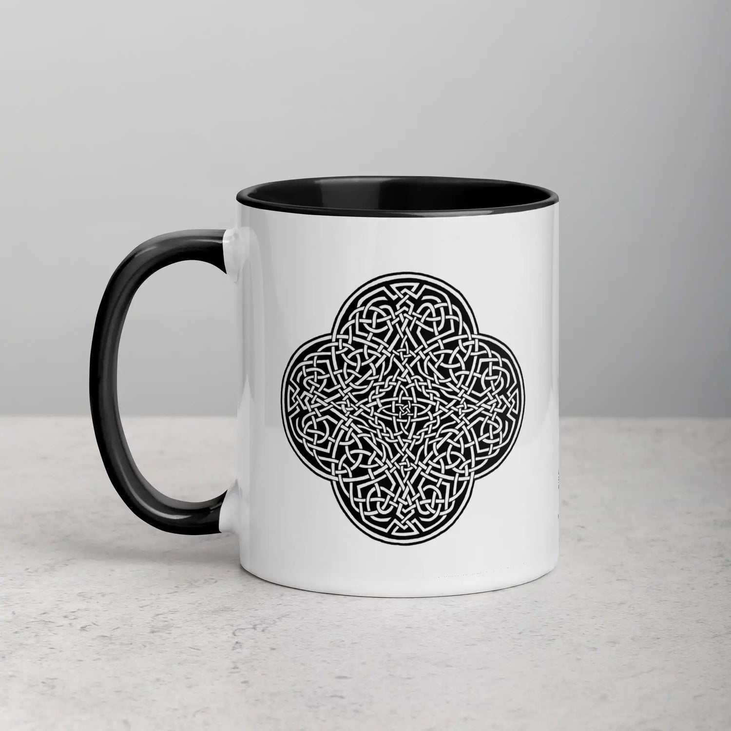 A tabletop displaying a Xigfireon white ceramic 11oz mug featuring the Black & White Solid iteration of the Reach Of The Spirit Celtic knot design, with black handle and inside. This Celtic knot symbolizes Mother Earth.