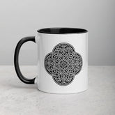 A tabletop displaying a Xigfireon white ceramic 11oz mug featuring the Black & White Solid iteration of the Reach Of The Spirit Celtic knot design, with black handle and inside. This Celtic knot symbolizes Mother Earth.