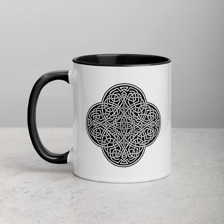 A tabletop displaying a Xigfireon white ceramic 11oz mug featuring the Black & White Solid iteration of the Reach Of The Spirit Celtic knot design, with black handle and inside. This Celtic knot symbolizes Mother Earth.