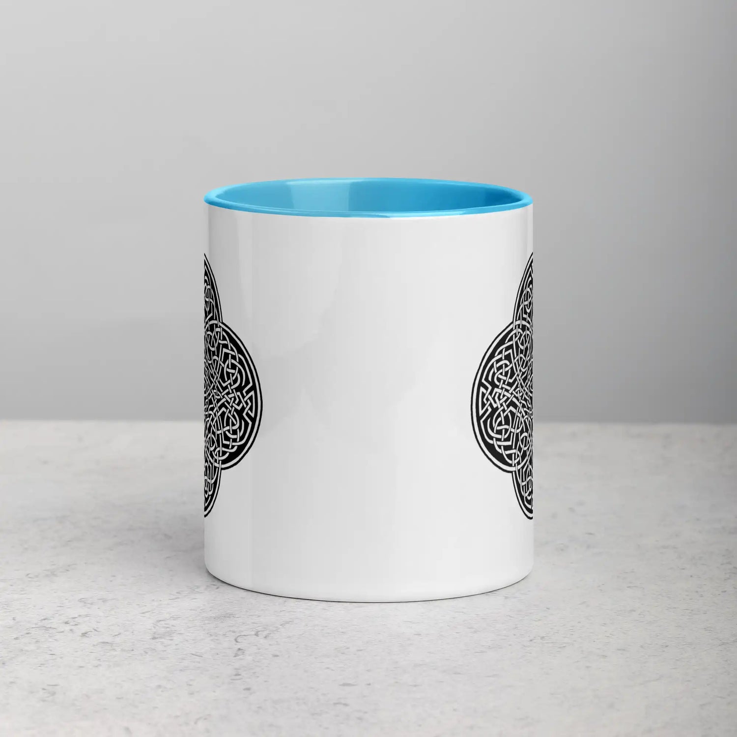 A tabletop displaying a Xigfireon white ceramic 11oz mug featuring the Black & White Solid iteration of the Reach Of The Spirit Celtic knot design, with blue handle and inside. This Celtic knot symbolizes Mother Earth.