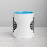 A tabletop displaying a Xigfireon white ceramic 11oz mug featuring the Black & White Solid iteration of the Reach Of The Spirit Celtic knot design, with blue handle and inside. This Celtic knot symbolizes Mother Earth.