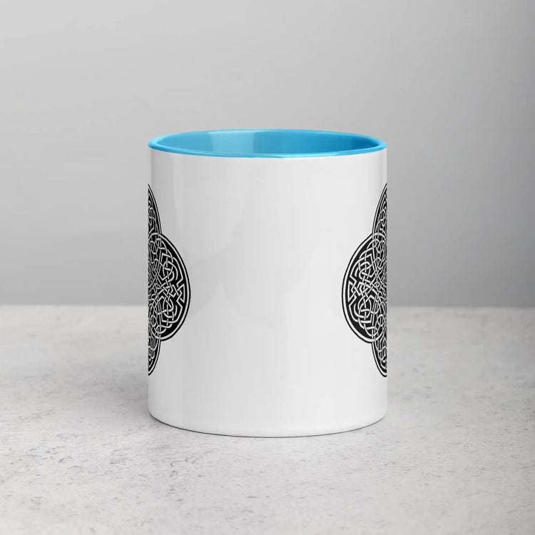 A tabletop displaying a Xigfireon white ceramic 11oz mug featuring the Black & White Solid iteration of the Reach Of The Spirit Celtic knot design, with blue handle and inside. This Celtic knot symbolizes Mother Earth.