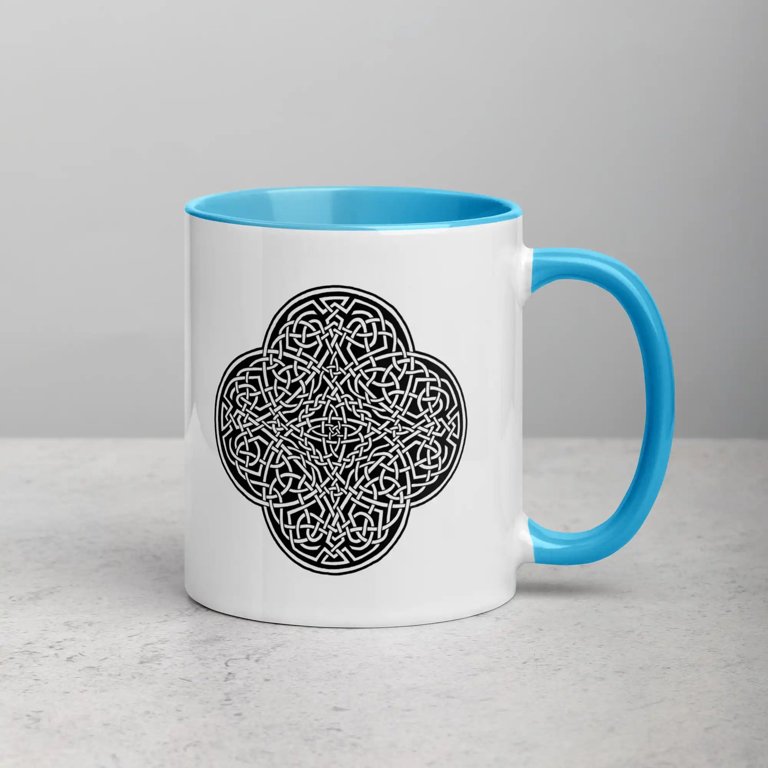 A tabletop displaying a Xigfireon white ceramic 11oz mug featuring the Black & White Solid iteration of the Reach Of The Spirit Celtic knot design, with blue handle and inside. This Celtic knot symbolizes Mother Earth.