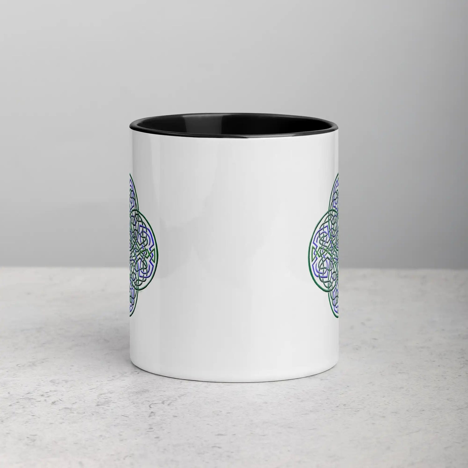 A tabletop displaying a Xigfireon white ceramic 11oz mug featuring the Living Colour iteration of the Reach Of The Spirit Celtic knot design, with black handle and inside. This Celtic knot symbolizes Mother Earth.