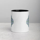 A tabletop displaying a Xigfireon white ceramic 11oz mug featuring the Living Colour iteration of the Reach Of The Spirit Celtic knot design, with black handle and inside. This Celtic knot symbolizes Mother Earth.