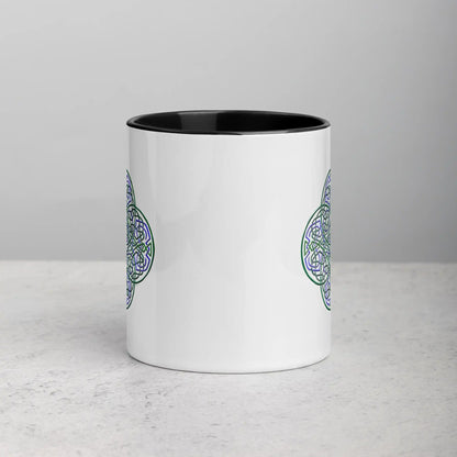 A tabletop displaying a Xigfireon white ceramic 11oz mug featuring the Living Colour iteration of the Reach Of The Spirit Celtic knot design, with black handle and inside. This Celtic knot symbolizes Mother Earth.