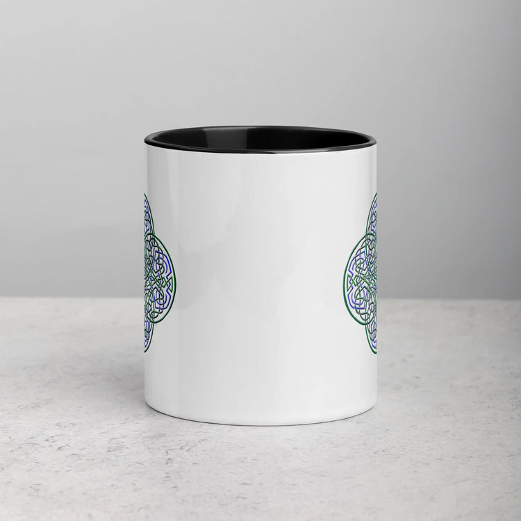 A tabletop displaying a Xigfireon white ceramic 11oz mug featuring the Living Colour iteration of the Reach Of The Spirit Celtic knot design, with black handle and inside. This Celtic knot symbolizes Mother Earth.
