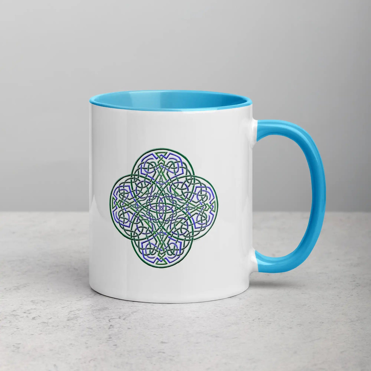 A tabletop displaying a Xigfireon white ceramic 11oz mug featuring the Living Colour iteration of the Reach Of The Spirit Celtic knot design, with blue handle and inside. This Celtic knot symbolizes Mother Earth.