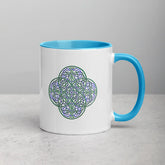 A tabletop displaying a Xigfireon white ceramic 11oz mug featuring the Living Colour iteration of the Reach Of The Spirit Celtic knot design, with blue handle and inside. This Celtic knot symbolizes Mother Earth.