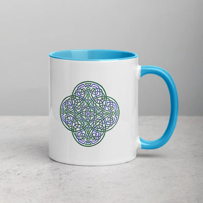 A tabletop displaying a Xigfireon white ceramic 11oz mug featuring the Living Colour iteration of the Reach Of The Spirit Celtic knot design, with blue handle and inside. This Celtic knot symbolizes Mother Earth.