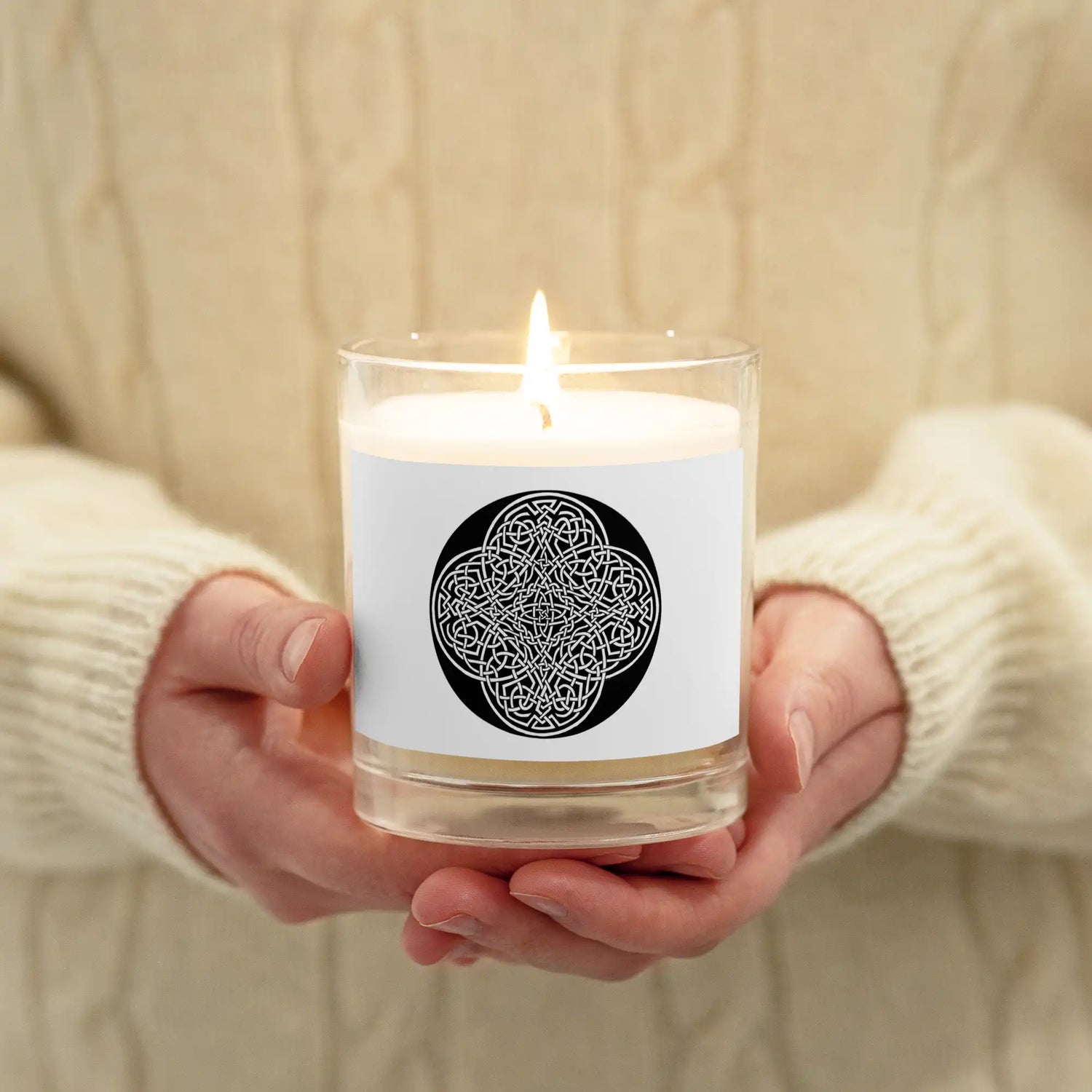 A person holding a Xigfireon glass jar soy wax candle featuring the Black & White Circle iteration of the Reach Of The Spirit Celtic knot design. The `Reach Of The Spirit` Celtic knot symbolizes Mother Earth.