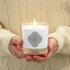 A person holding a Xigfireon glass jar soy wax candle featuring the Black & White Hollow iteration of the Reach Of The Spirit Celtic knot design. The Reach Of The Spirit Celtic knot symbolizes Mother Earth.