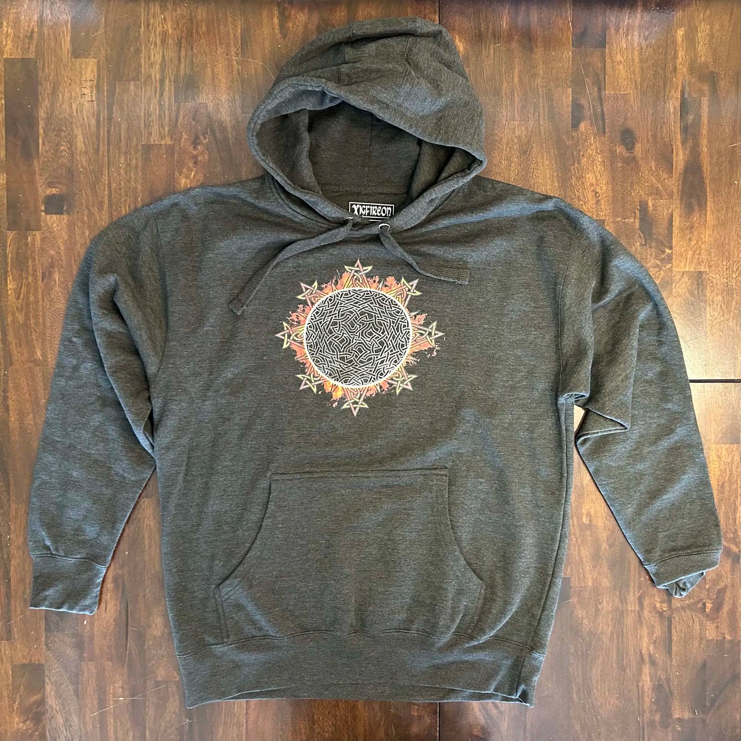 A wooden tabletop with a charcoal heather Xigfireon graphic hoodie featuring the Eclipsed iteration of the Morning Star Fire Celtic knot design, inspired by the 2023 Annular Solar Eclipse.