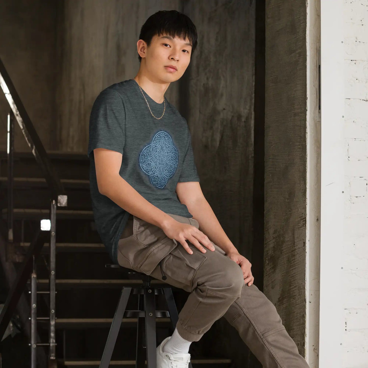 A young man sitting on a stool wearing a dark heather graphic t-shirt featuring the Blue Slate iteration of the Reach Of The Spirit Celtic knot design.