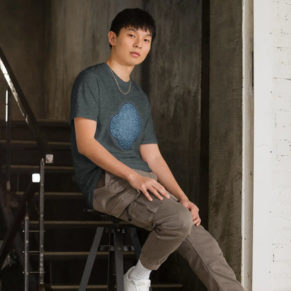 A young man sitting on a stool wearing a dark heather graphic t-shirt featuring the Blue Slate iteration of the Reach Of The Spirit Celtic knot design.